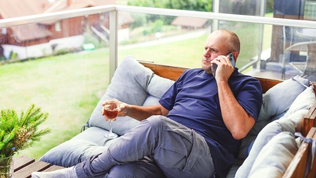Middle-aged man sitting on balcony, talking on phone and holding a glass of wine.