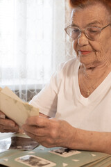 An elderly woman is sitting at a table, looking at old photographs. She is wearing a white T-shirt and glasses. Her red hair is tied in a bun.
In front of her is an old photo album with yellowed pages