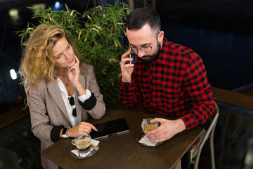 Casually dressed man and woman enjoying coffee in a cafe setting, engaged in conversation, with the man attentively on a call, creating a warm and relaxed atmosphere