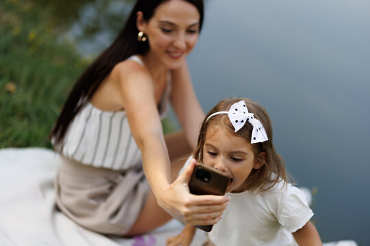 Mother and daughter enjoying time together while taking selfies outdoors by a tranquil water backdrop, portraying joy and bonding