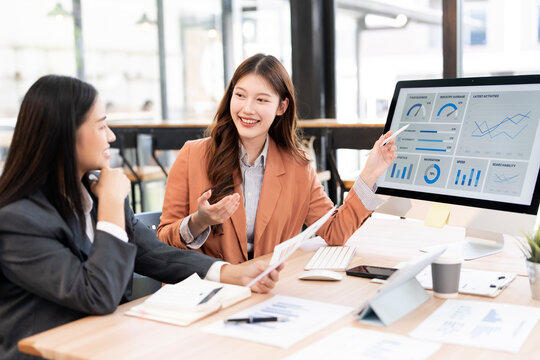 Two happy young Asian businesswomen discussing a business plan, pointing at data charts on a computer screen and collaborating in a modern office meeting. - Powered by Adobe
