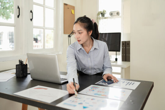 A businesswoman in a modern office setting is focused while working on a laptop and reviewing documents at her desk, analyzing data.