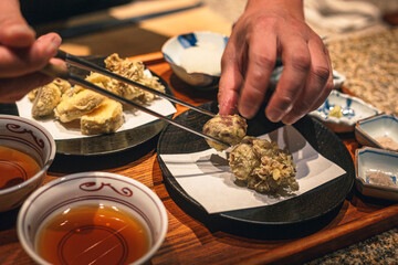 Close-up of hands preparing vegetable tempura with chopsticks, served with dipping sauce in traditional Japanese bowls on a wooden tray.