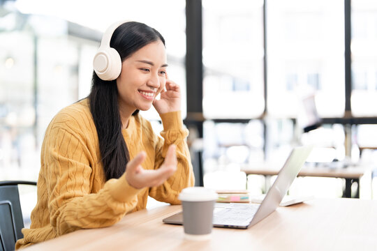 A happy young Asian woman in casual attire smiles while listening on wireless headphones and using a laptop in a modern cafe.