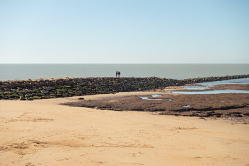 Three people venture into the sea along a path of stones and rocks over the water. View of the corrales of Chipiona. Three people walking over traditional fishing traps. View of a beach.