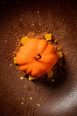 A vibrant pumpkin-shaped dessert sits elegantly on a textured brown plate, garnished artistically