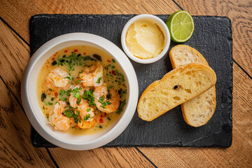 A flavorful shrimp dish sits in a bowl with herbs, accompanied by butter, lime, and golden toast