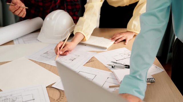 Architect and safety engineer discussion about building blueprint displaying on laptop on table with architectural document and wooden block scatter around at modern office. Closeup. Delineation.