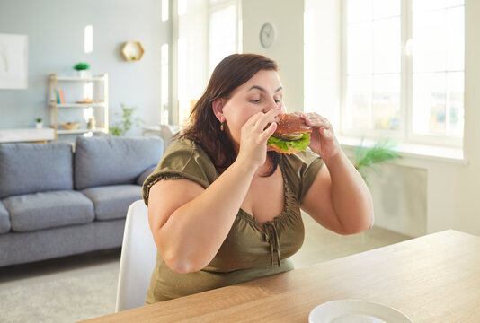 Obese hungry woman with excessive fat eating unhealthy fast food, tasty hamburger, juicy burger, cheeseburger, chubby overweight plus size big fat fit lady with good appetite enjoys excess calories