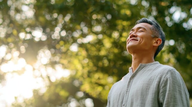 Medium view of a healthy Asian adult outdoors, standing in nature, closing eyes while taking a deep breath of fresh air, smiling with a calm and energized expression, posture open and relaxed
