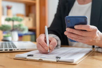 Woman's hands jotting numbers on a clipboard while holding a smartphone, working through budgets and financial reports in a modern home office with laptop and paperwork nearby