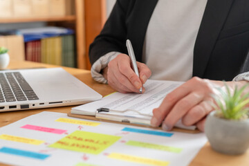 Business professional hands analyzing financial data on a clipboard, making notes while reviewing charts and reports on a desk with a laptop and potted plant