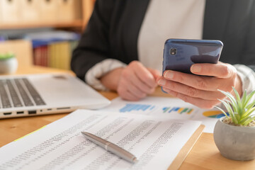 Businesswoman's hands holding a smartphone while checking financial data, reports, and charts on a desk with a laptop, emphasizing modern business analysis and technology integration