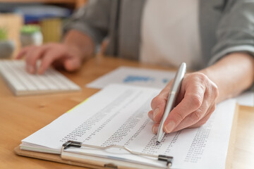 Person using a pen to review financial numbers on a clipboard, while simultaneously operating a computer keyboard, focusing on data analysis and strategic planning