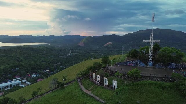 Rightward linear move along Mount Tapyas summit reveals the steel cross, CORON hillside letters, and sweeping green valleys and bays under a calm sunset sky above Coron, Palawan.