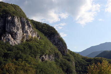 Rocky karst landscape of the Dinaric Alps in Bosnia and Herzegovina. Steep limestone cliffs rise above densely forested slopes, with rolling mountain ridges in the distance under a partly cloudy sky.