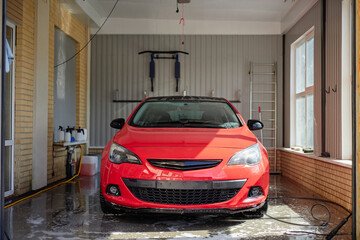 Obraz premium Red car parked inside car wash bay with wet floor reflecting vehicle, sunlight streaming through large windows, cleaning equipment and tools mounted on walls in background