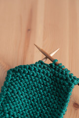 Close-up of green knitting fabric with wooden needles on light wooden table. Vertical minimalist image with neutral background and copy space. Concept of handmade crafts and cozy lifestyle