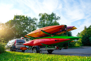 Colorful kayaks canoes securely attached to a trailer being pulled by car. Trailer is parked on asphalt near a grassy area and green trees, suggesting a park or recreation area