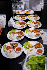 Plates of Indonesian fried chicken with rice, sambal, cucumber, and lettuce arranged neatly on a black counter in restaurant kitchen, ready to be served.