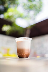 A glass of iced chocolate with milk layers placed on a table in outdoor cafe setting with soft bokeh background and bright daylight.