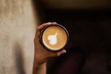 Hand holding a cup of hot latte with heart-shaped latte art against a warm wall background, showcasing cozy cafe mood.