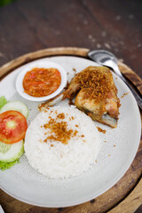 Indonesian fried chicken with steamed rice, sambal chili sauce, and fresh vegetables served on a rustic wooden table. Traditional Asian lunch dish.