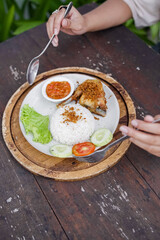 Indonesian fried chicken with steamed rice, sambal chili sauce, and fresh vegetables served on a rustic wooden table. Traditional Asian lunch dish.