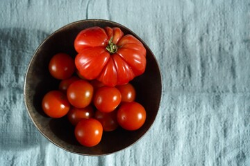 Overhead view of a bowl containing mixed tomatoes, featuring a large heirloom tomato among smaller cherry tomatoes, set against a textured blue fabric background for contrast.