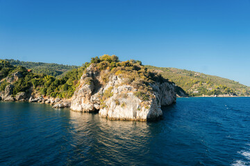 Scenic view of whitewashed houses with terracotta roofs built on a rocky cliff above the blue Aegean Sea in Skiathos, Greece. Mediterranean trees and clear turquoise water create a picturesque summer