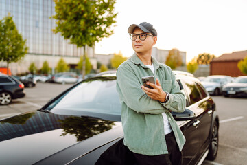 A young man wearing glasses and holding a phone stands next to a car in a parking lot. The man enjoys the sunny day with his phone next to a dark car. Conceptual image: transportation, leisure.