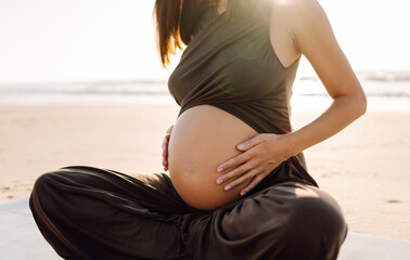 A pregnant woman meditates on beach in sun's rays. A beautiful expectant mother performs yoga on yoga mat at sunrise. Concepts of health, motherhood, an active lifestyle.