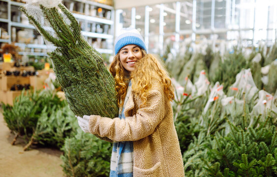 Smiling woman chooses a Christmas tree at busy market. A beautiful woman with bright scarf enjoys the New Year's atmosphere and holds Christmas tree. Concepts of holiday, shopping, and consumerism.