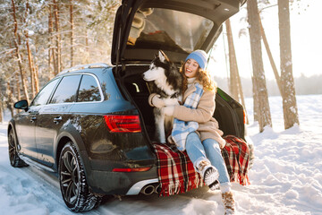 A cheerful woman sits in trunk of a car in snowy forest with her pet. A woman and her Siberian husky are on a road trip on a sunny winter day. Concepts include adventure, relaxation, and weekends.