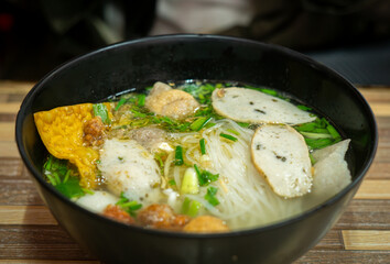 Close-up of a delicious Thai noodle soup served in a black bowl. The dish includes rice noodles, fish balls, sliced fish cakes, crispy wonton, fresh herbs, and clear flavorful broth. Authentic Asian s