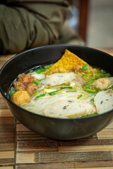 Close-up of a delicious Thai noodle soup served in a black bowl. The dish includes rice noodles, fish balls, sliced fish cakes, crispy wonton, fresh herbs, and clear flavorful broth. Authentic Asian s