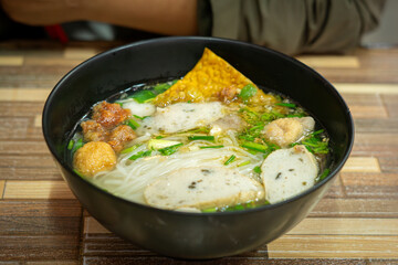 Close-up of a delicious Thai noodle soup served in a black bowl. The dish includes rice noodles, fish balls, sliced fish cakes, crispy wonton, fresh herbs, and clear flavorful broth. Authentic Asian s
