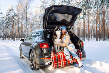 A cheerful woman sits in trunk of a car in snowy forest with her pet. A woman and her Siberian husky are on a road trip on a sunny winter day. Concepts include adventure, relaxation, and weekends.