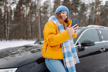 Portrait of happy woman in a bright jacket taking selfie while standing next to car on winter day. Beautiful woman with phone in a snowy forest, enjoying nature. Concept of transportation, travel.