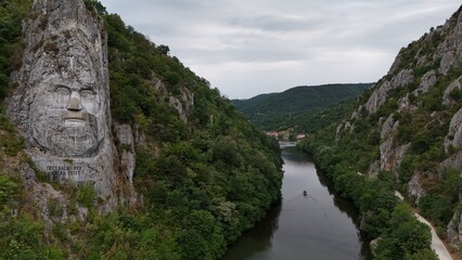 Decebalus Rex Overlooking the Danube's Dramatic Gorges. An expansive aerial or elevated view capturing the majestic Decebalus Rex rock sculpture on the left, carved into a colossal cliff face.