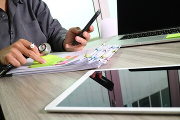 Doctor multitasking with smartphone, medical files, and laptop on office desk
