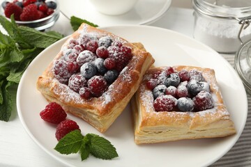 Tasty puff pastries with berries, powdered sugar and mint on white table, closeup