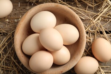 Raw chicken eggs in bowl and straw on wooden table, flat lay