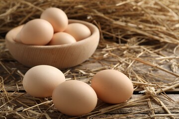 Raw chicken eggs in bowl and straw on wooden table, closeup