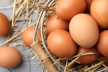 Raw chicken eggs, metal basket and straw on grey wooden table, flat lay