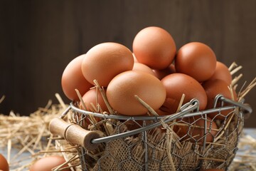 Raw chicken eggs, metal basket and straw on grey table against brown background, closeup