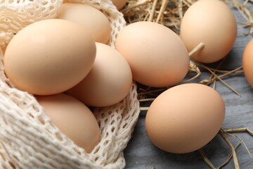 Raw chicken eggs, mesh bag and straw on grey wooden table, closeup