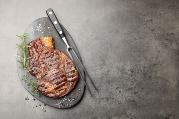 Delicious grilled beef steak with herbs, spices and carving fork on grey textured table, top view. Space for text