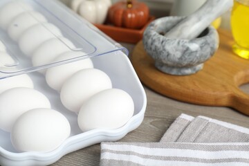 Raw chicken eggs in plastic container and kitchen utensils on wooden countertop, closeup