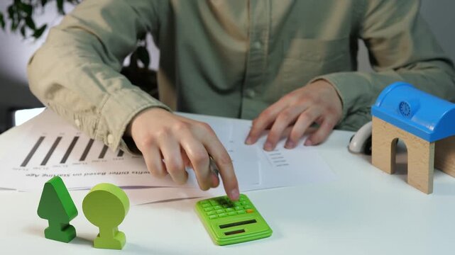 Focused hands engage in a complex calculation for a development project, referencing analytical graphs while surrounded by symbolic representations of housing and ecology. T